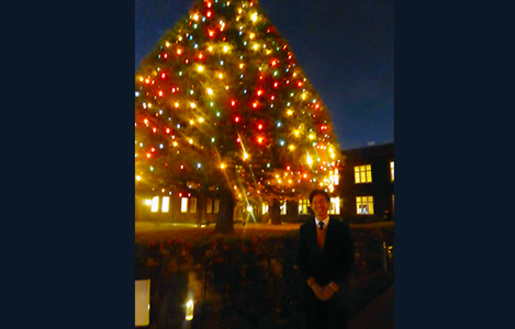 立教大学の同窓会『中野立教会　クリスマス会』夜になってまずます輝いて見える電飾が美しい2階建ての建物よりもずっと大きな木の前に立つ笑顔の石坂わたる